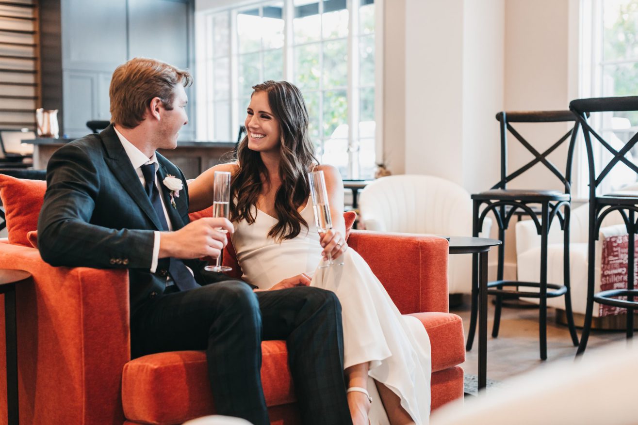 A bride and groom sit on a sofa smiling at each other. There are large windows behind them.