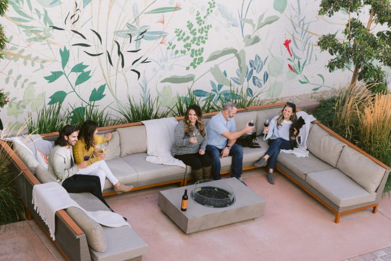 5 people sitting on u-shaped patio furniture around a fire pit drinking wine. One woman is holding a small black dog.